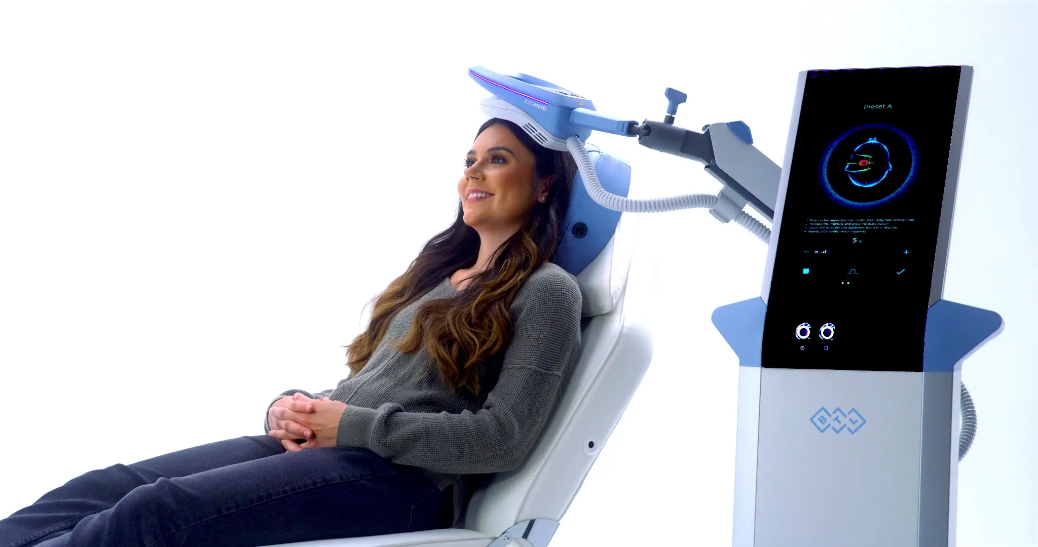EXOMIND treatment demonstration with a woman seated beside the system and the applicator positioned above her head.